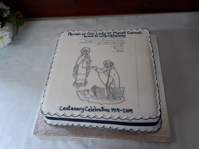 A decorated cake saying 'Centenary Celebration 1919-2019' and 'Parish of Our Lady of Mount Carmel and St Wilfrid' and decorated in blue, with an outline image of the stone relief on the church, depicting Our Lady, the infant Christ and St Wilfrid