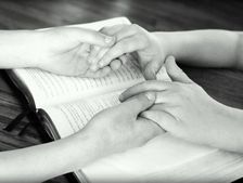 Two people's hands clasped over an open Bible; praying together