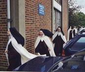 Carmelite nuns in dark habits and white cloaks, walking down the side of St Richard's Chapel, cloaks blowing in the breeze.