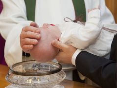 A baby is held over a glass bowl and baptised by a priest